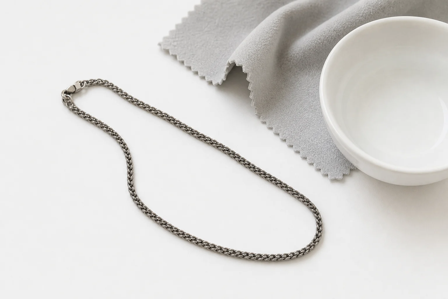 Is Titanium Safe for Sensitive Skin? What Actually Makes It Different (And Why It Won’t Tarnish) 6 Titanium necklace chain on white surface with soft microfiber cloth and water bowl showing minimal cleaning setup for titanium jewelry care
